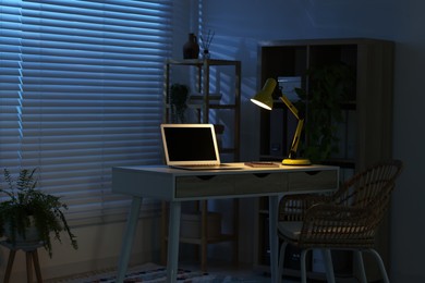 Photo of Comfortable workplace with laptop and lamp on desk at home office in evening