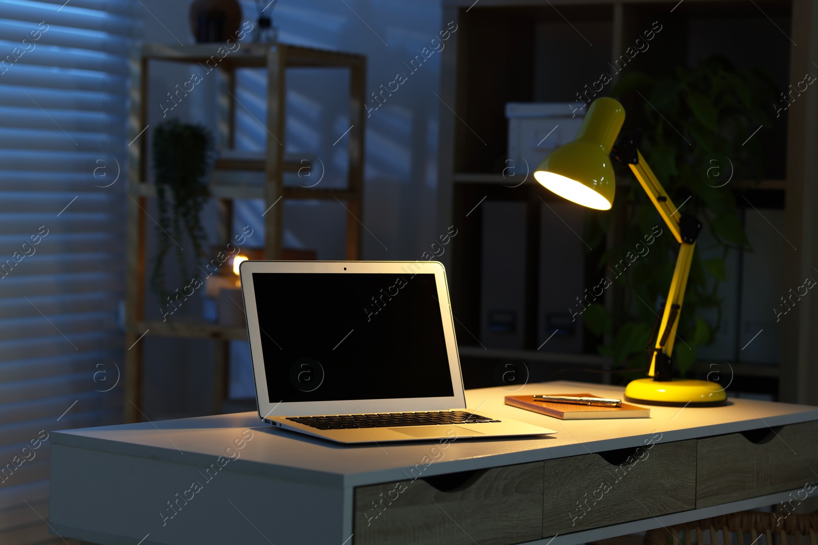 Comfortable workplace with laptop and lamp on desk at home office in evening Photo of Comfortable workplace with laptop and lamp on desk at home office in evening