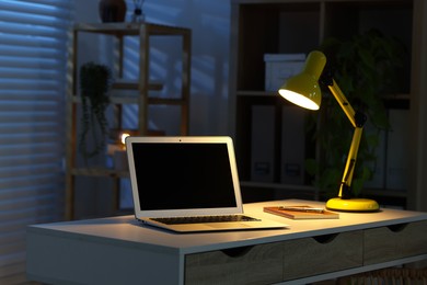 Photo of Comfortable workplace with laptop and lamp on desk at home office in evening