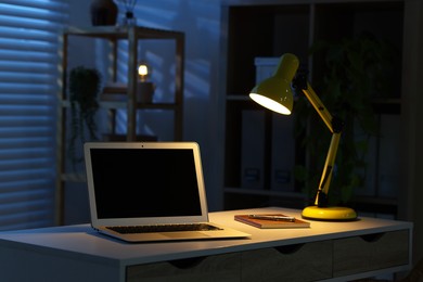 Photo of Comfortable workplace with laptop and lamp on desk at home office in evening