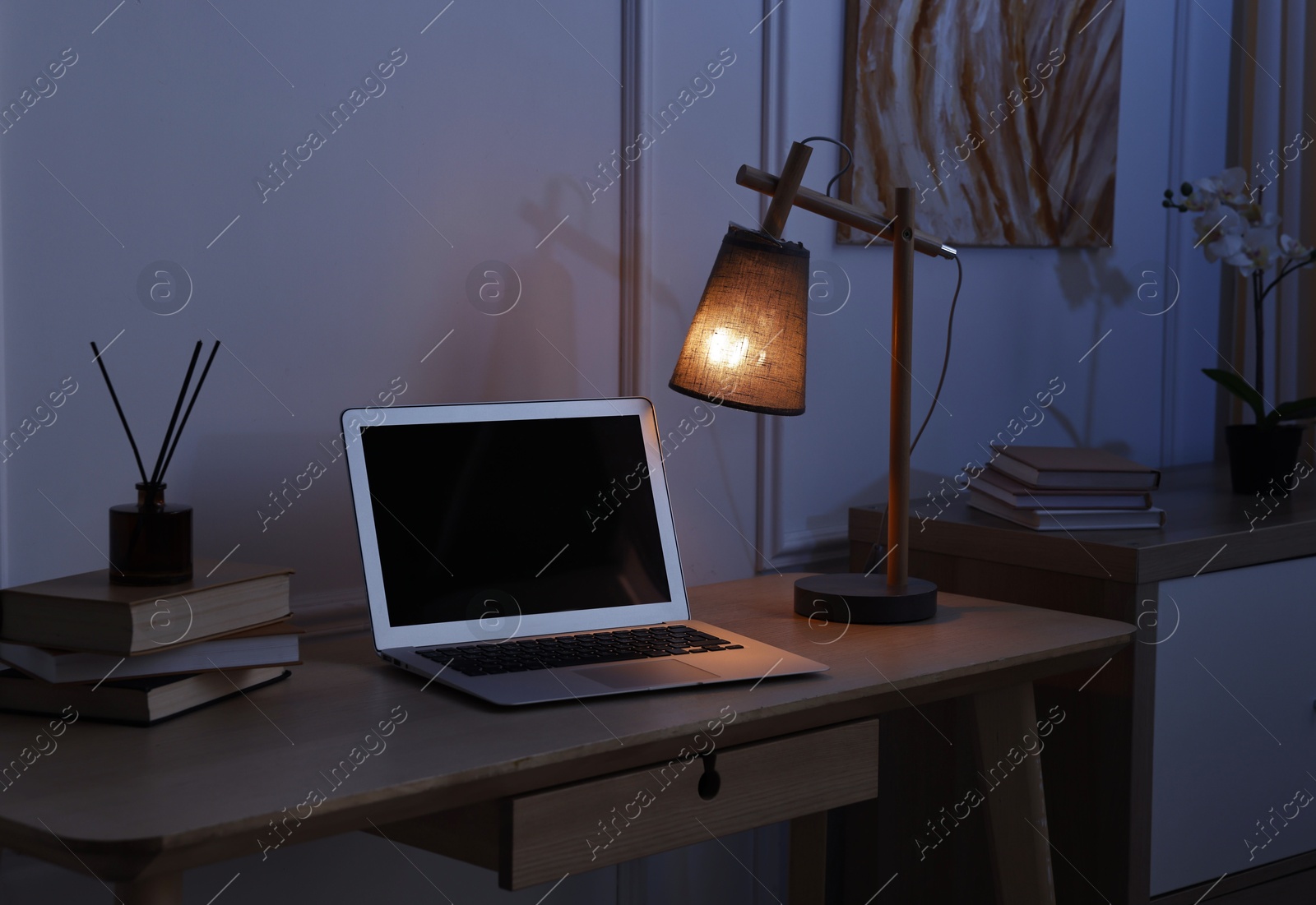 Comfortable workplace with modern laptop and lamp on wooden desk at home office in evening Photo of Comfortable workplace with modern laptop and lamp on wooden desk at home office in evening