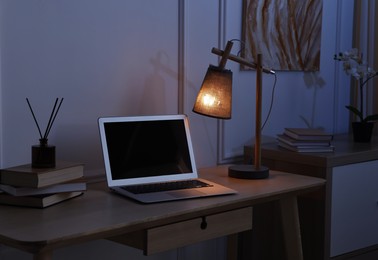 Photo of Comfortable workplace with modern laptop and lamp on wooden desk at home office in evening