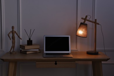 Photo of Comfortable workplace with modern laptop and lamp on wooden desk at home office in evening