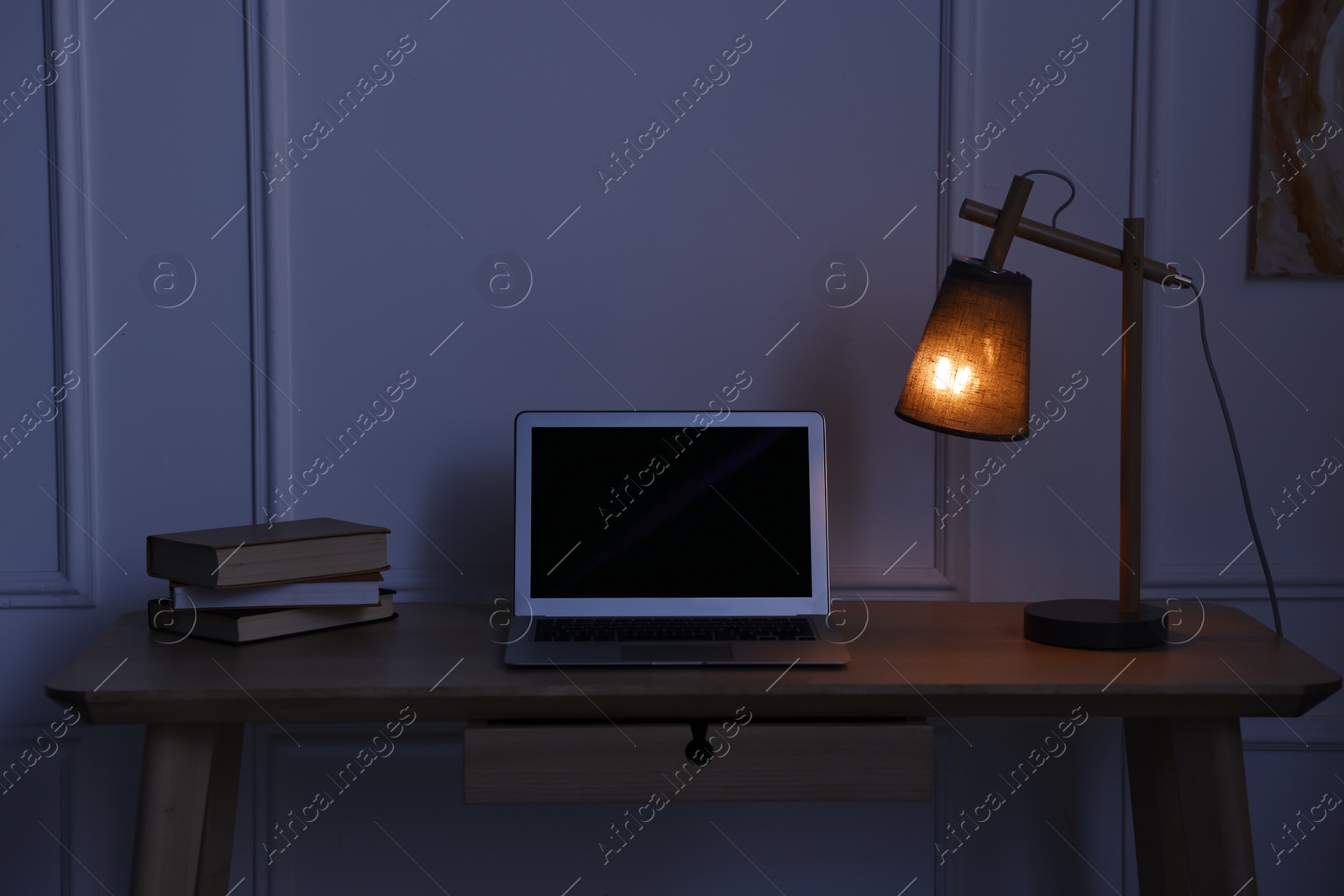 Comfortable workplace with modern laptop and lamp on wooden desk at home office in evening Photo of Comfortable workplace with modern laptop and lamp on wooden desk at home office in evening