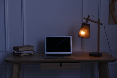 Photo of Comfortable workplace with modern laptop and lamp on wooden desk at home office in evening