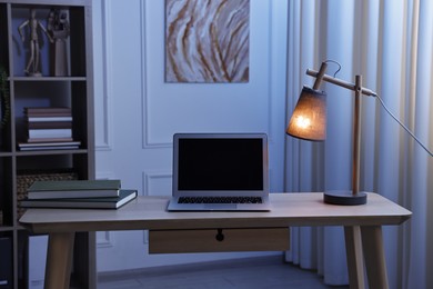 Photo of Comfortable workplace with modern laptop and lamp on wooden desk at home office in evening