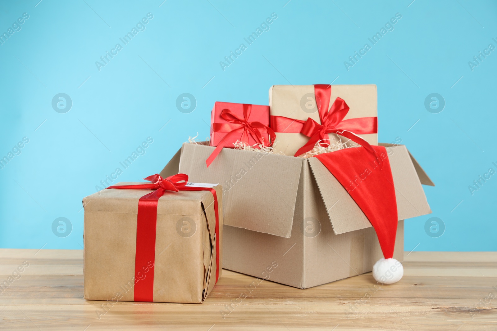 Parcel with gift boxes and Santa hat on wooden table against light blue background Photo of Parcel with gift boxes and Santa hat on wooden table against light blue background