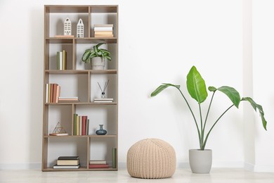 Photo of Wooden shelving unit with different home decor, pouf and houseplant near white wall indoors