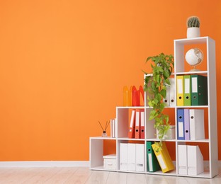 Photo of Shelving unit with folders and plants near orange wall indoors. Space for text