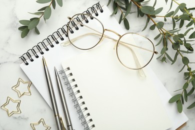 Paper clips, notebooks, pens, glasses and green branches on white marble table, flat lay