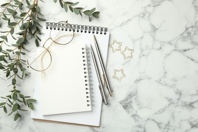 Paper clips, notebooks, pens, glasses and green branches on white marble table, flat lay. Space for text