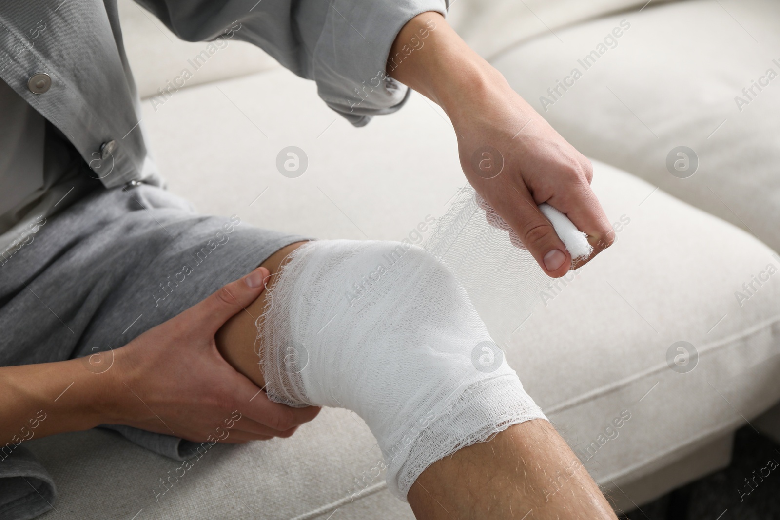 Photo of Man wrapping knee in medical bandage on sofa at home, closeup