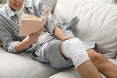 Photo of Man with medical bandage on his knee reading at home, closeup