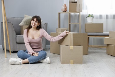 Photo of Moving day. Happy woman sitting on floor near cardboard boxes in her new home