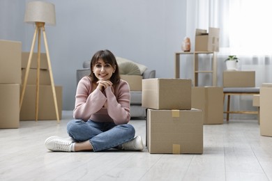 Photo of Moving day. Happy woman sitting on floor near cardboard boxes in her new home