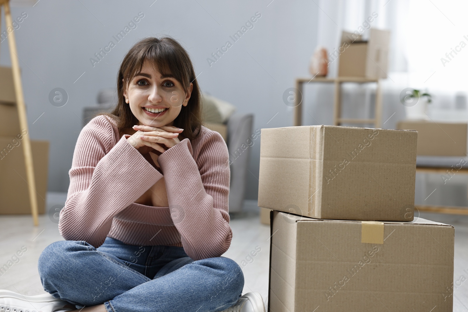 Photo of Moving day. Happy woman sitting on floor near cardboard boxes in her new home