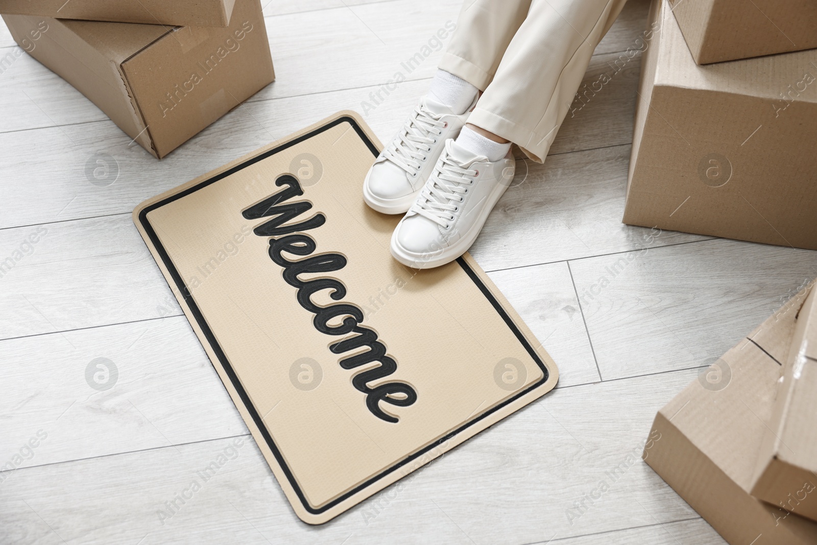 Photo of Moving day. Woman sitting near cardboard boxes and doormat with word Welcome on floor in her new home, closeup