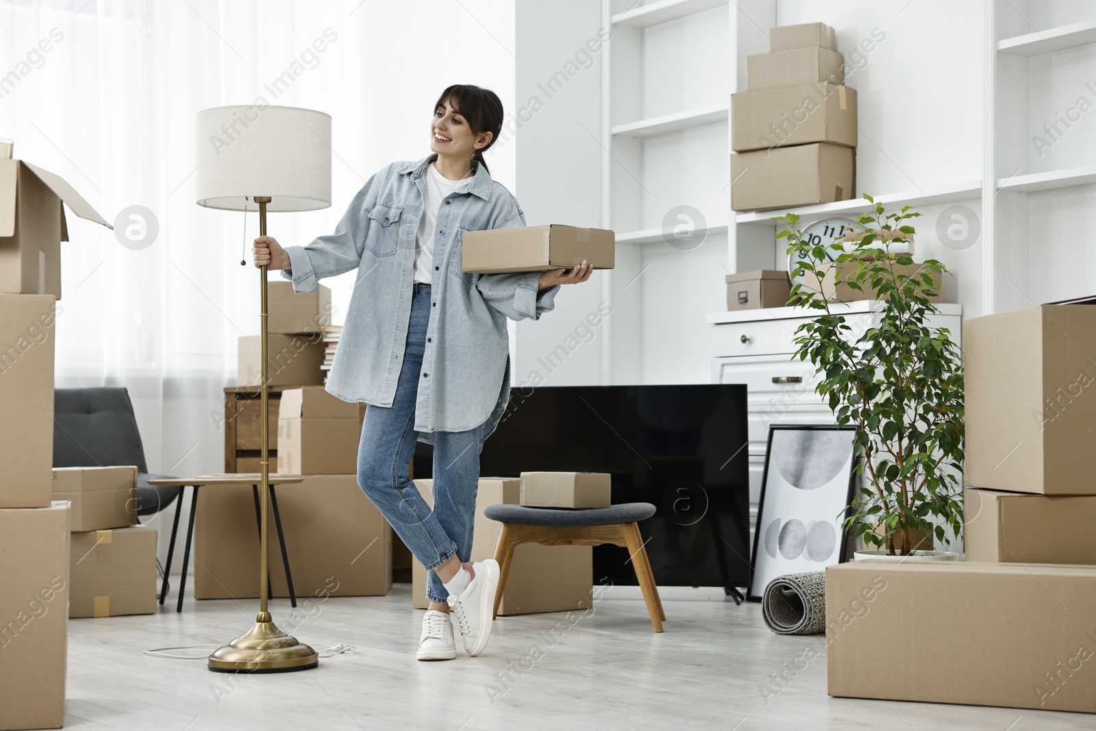 Photo of Moving day. Happy woman with lamp and cardboard box in her new home