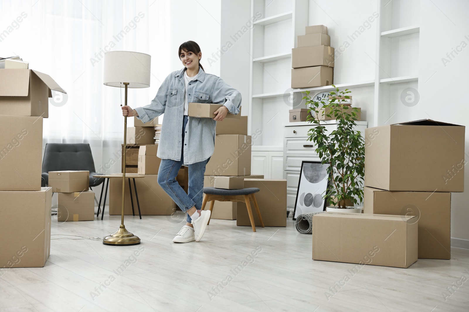 Photo of Moving day. Happy woman with lamp and cardboard box in her new home