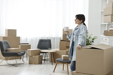 Photo of Moving day. Happy woman with cup of drink and cardboard boxes in her new home