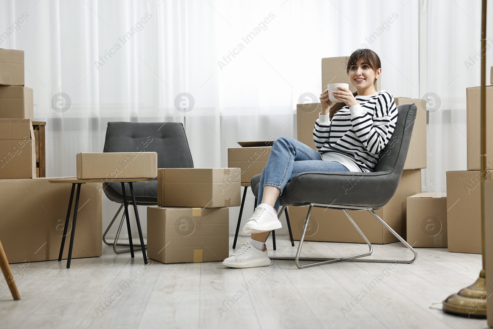 Photo of Moving day. Happy woman with cup of drink and cardboard boxes in new home