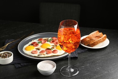 Photo of Tasty brunch. Aperol cocktail, delicious fried eggs with sausage served on black table, closeup