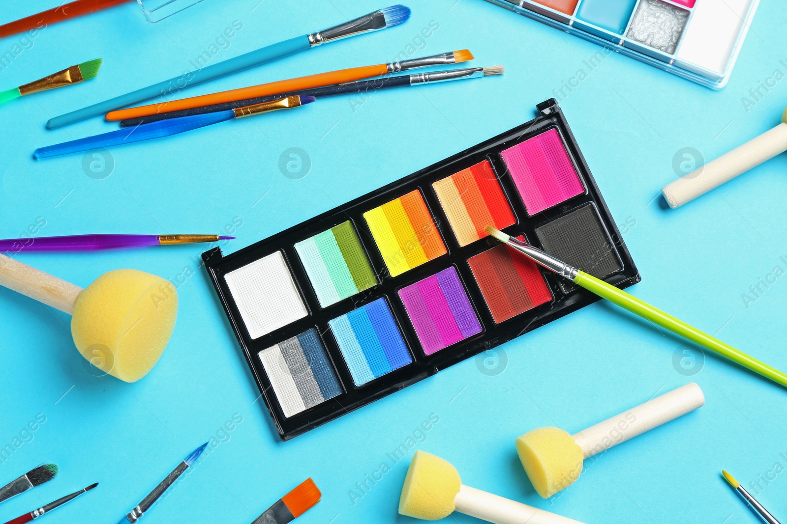Bright face paints, brushes and sponges on light blue background, flat lay Photo of Bright face paints, brushes and sponges on light blue background, flat lay