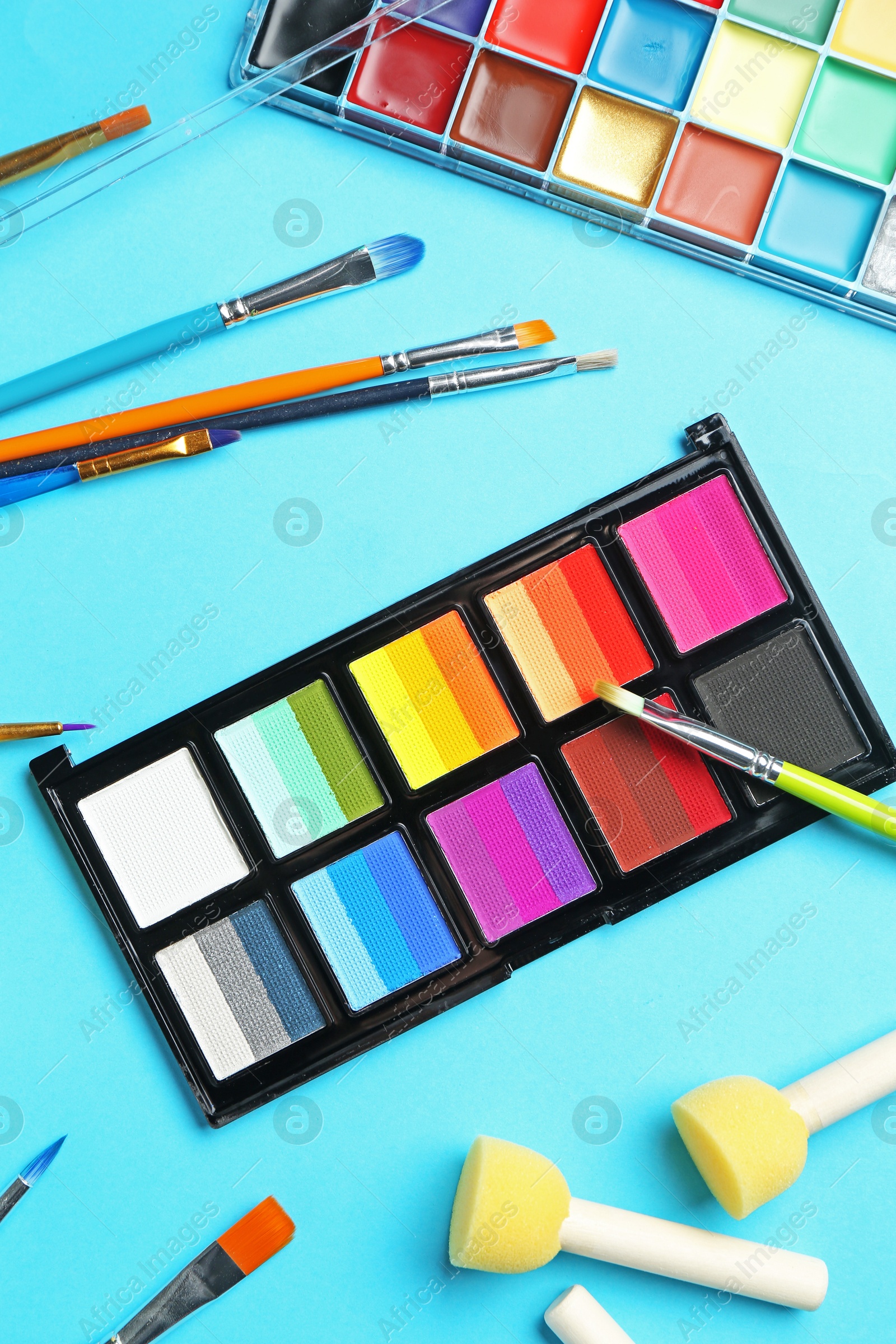 Bright face paints, brushes and sponges on light blue background, flat lay Photo of Bright face paints, brushes and sponges on light blue background, flat lay