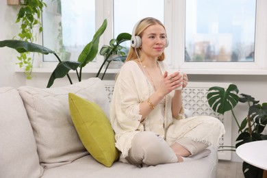 Photo of Feng shui. Young woman with headphones and cup of drink meditating on couch at home