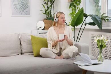 Photo of Feng shui. Young woman with headphones and cup of drink meditating on couch at home