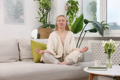 Photo of Feng shui. Young woman with headphones meditating on couch at home