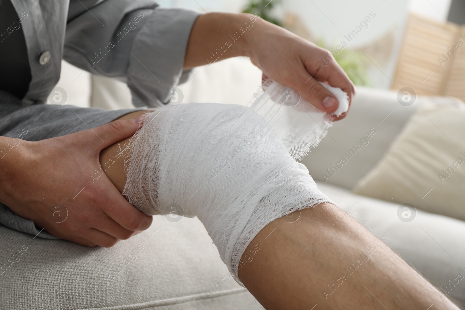 Photo of Man wrapping knee in medical bandage on sofa at home, closeup