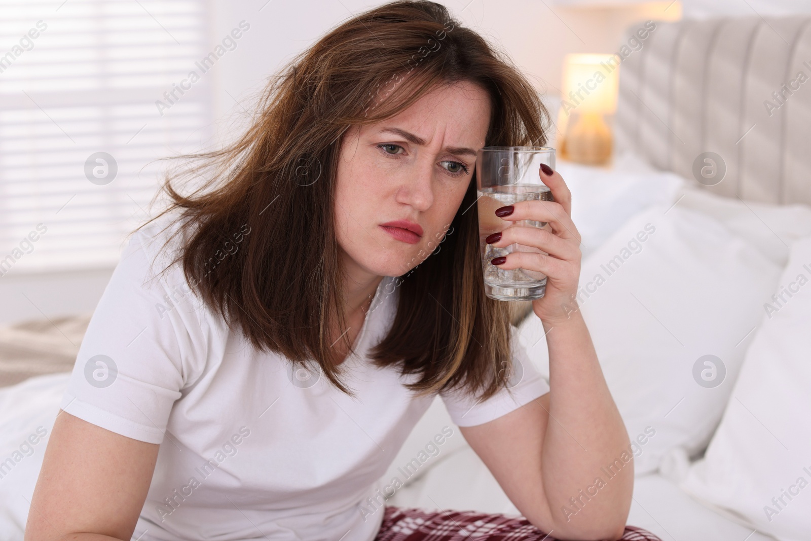 Photo of Unhappy woman with glass of water suffering from hangover on bed at home