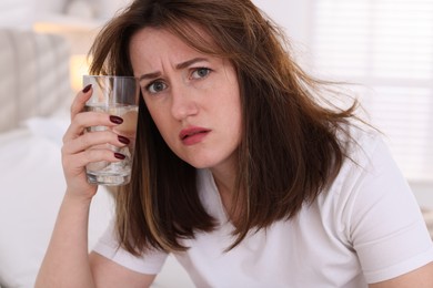 Unhappy woman with glass of water suffering from hangover on bed at home