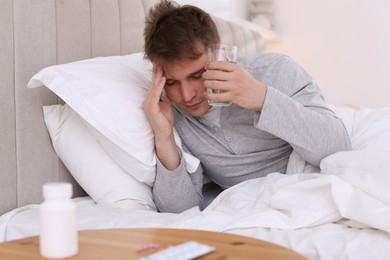 Unhappy young man with glass of water suffering from hangover on bed at home