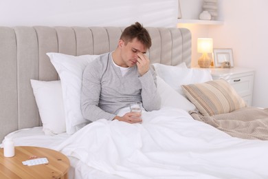 Photo of Unhappy young man with glass of water suffering from hangover on bed at home