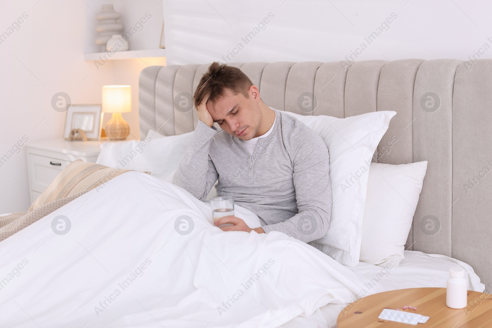 Photo of Unhappy young man with glass of water suffering from hangover on bed at home