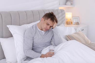 Photo of Unhappy young man with glass of water suffering from hangover on bed at home