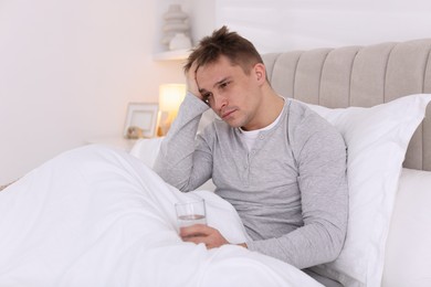 Photo of Unhappy young man with glass of water suffering from hangover on bed at home