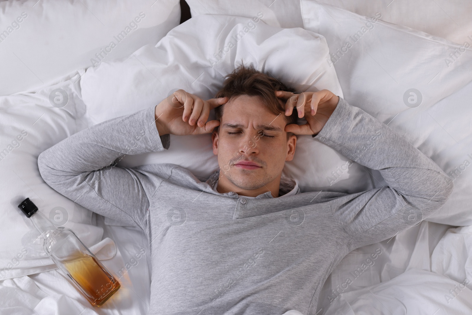 Photo of Unhappy young man with bottle of whiskey suffering from hangover on bed at home, top view