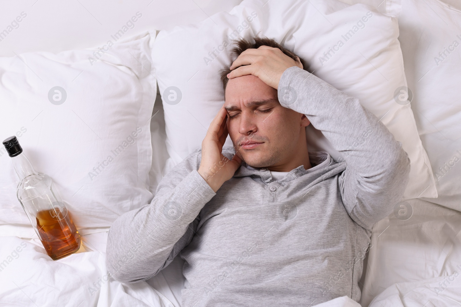 Photo of Unhappy young man with bottle of whiskey suffering from hangover on bed at home, top view