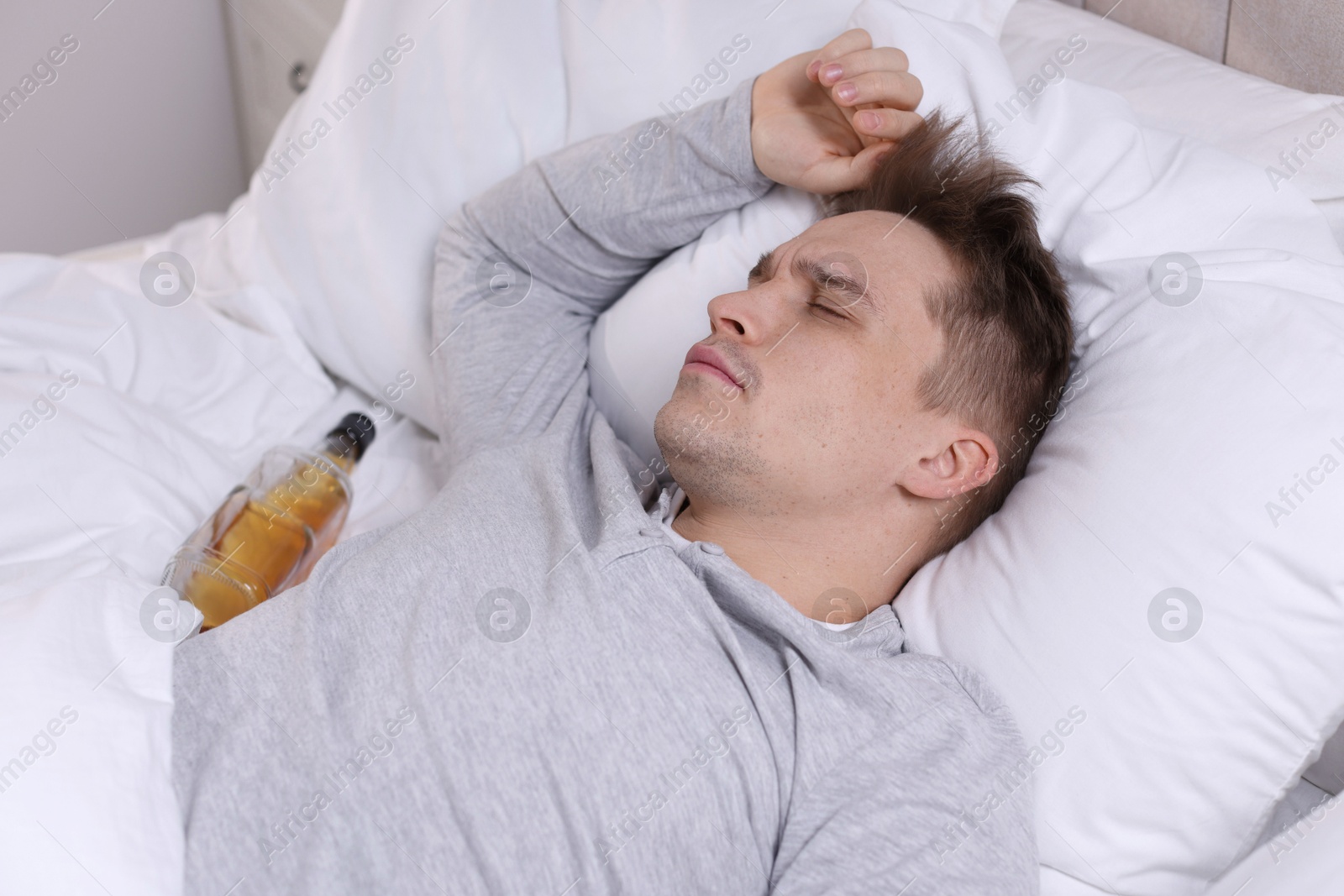 Photo of Unhappy young man with bottle of whiskey suffering from hangover on bed at home