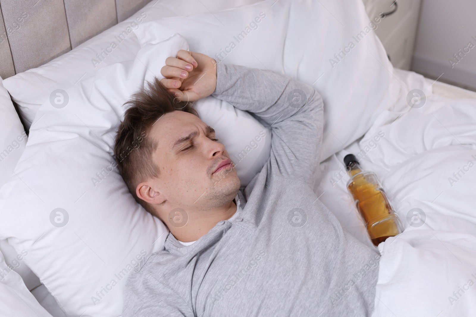 Photo of Unhappy young man with bottle of whiskey suffering from hangover on bed at home, above view