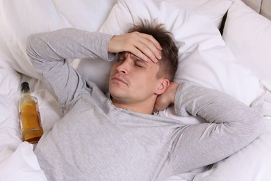 Photo of Unhappy young man with bottle of whiskey suffering from hangover on bed at home, above view