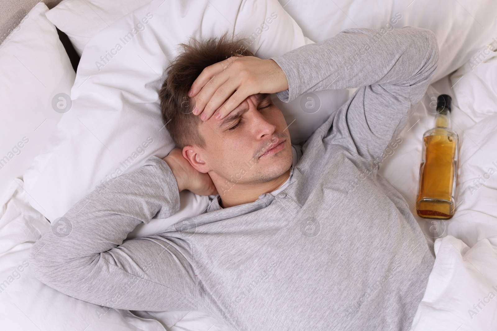 Photo of Unhappy young man with bottle of whiskey suffering from hangover on bed at home, top view