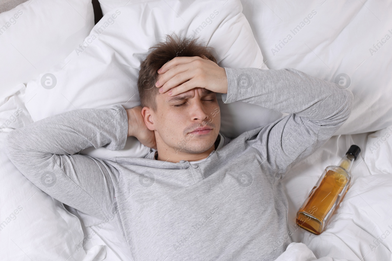 Photo of Unhappy young man with bottle of whiskey suffering from hangover on bed at home, top view