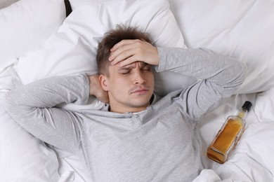 Unhappy young man with bottle of whiskey suffering from hangover on bed at home, top view