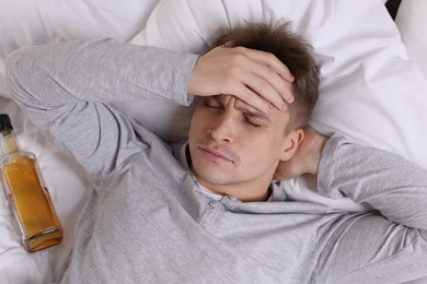 Unhappy young man with bottle of whiskey suffering from hangover on bed at home, top view