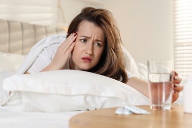 Photo of Unhappy woman with glass of water and pills suffering from hangover on bed at home