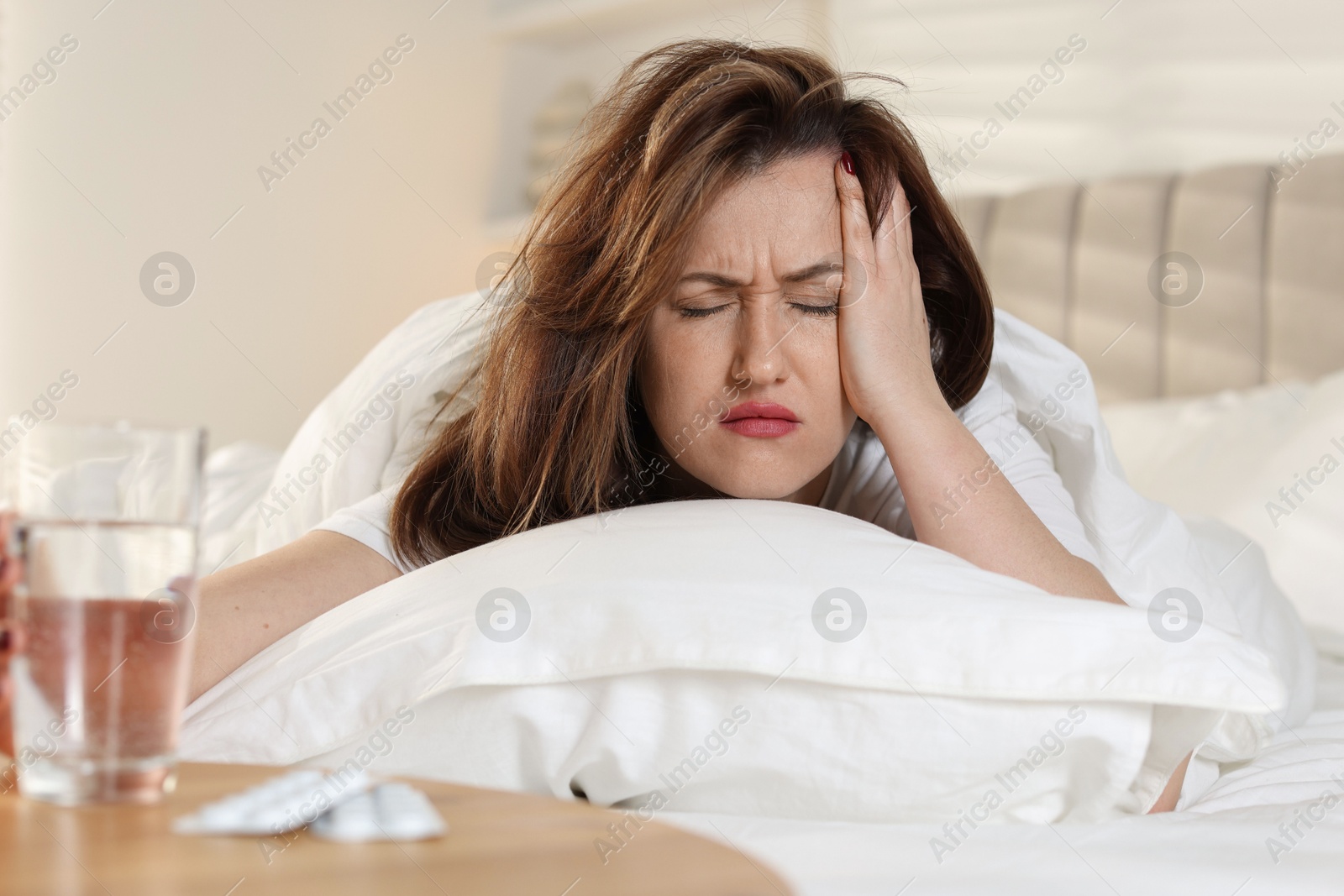 Photo of Unhappy woman with glass of water and pills suffering from hangover on bed at home